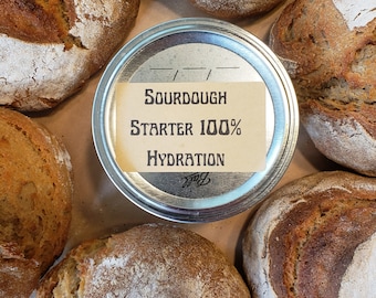 May include: Overhead shot of several round loaves of sourdough bread surrounding a jar lid. The jar lid has a label that reads "Sourdough Starter 100% Hydration". The bread has a crusty exterior and a light dusting of flour.