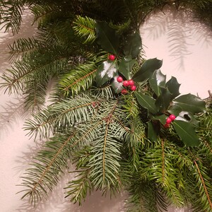 May include: A close-up of a Christmas wreath made with evergreen branches and holly sprigs with red berries.