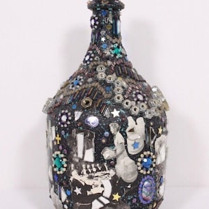 May include: A decorative bottle covered in a mosaic of beads, sequins, and metal hardware. The dark bottle features a collage of black and white images and is adorned with blue, silver, and turquoise accents. The bottle's surface is textured with various embellishments.