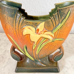 May include: A ceramic vase with a floral design. The vase features a yellow lily with green leaves against an orange background. The vase has a square base and curved handles. The top edge is scalloped, and the vase is approximately 8 inches tall.