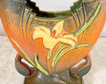 May include: A ceramic vase with a floral design. The vase features a yellow lily with green leaves against an orange background. The vase has a square base and curved handles. The top edge is scalloped, and the vase is approximately 8 inches tall.