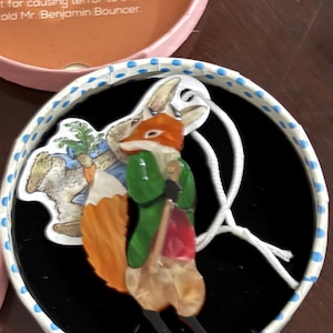 May include: A fox-shaped brooch featuring a character in a green jacket and red vest, holding a walking stick. The brooch is displayed in a round box with a lid labeled MR. TOD and text describing a fox causing terror to the Bouncer family.