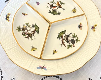 May include: A two-tiered serving dish set. The bottom tier is a large, round, off-white plate with a textured rim. The top tier is a three-sectioned plate with gold trim, featuring bird and butterfly illustrations. The plates are on a white tablecloth.