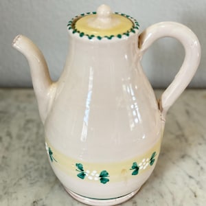 May include: A ceramic teapot with a light pink body, yellow lid, and a curved handle. The lid and base feature green and white floral designs. The spout is slightly chipped. The teapot is likely used for serving tea.
