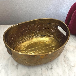 May include: Oval-shaped, antique brass bowl with ornate floral and leaf patterns around the rim. The bowl has a hammered texture on the interior and small handles on each side. The bowl is a warm gold color.