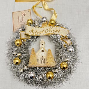 May include: A Christmas wreath with silver tinsel, gold and silver ornaments, and a banner reading "Silent Night." A small church and trees are in the center. A gold ribbon is at the top.