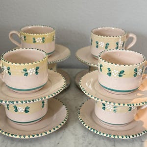 May include: Set of four ceramic teacups and saucers. The cups and saucers are pale pink with a yellow band featuring white flowers and green leaves. The rims are scalloped and trimmed with green.