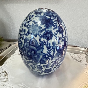 May include: A large, white ceramic egg with a blue and white floral pattern. The egg is sitting on a white lace doily.