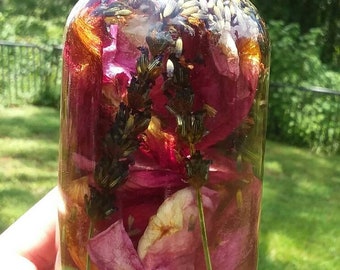 Op de afbeelding: Een heldere glazen fles gevuld met een mengsel van gedroogde rozenblaadjes en lavendelbloemen. De fles heeft een zilveren deksel.