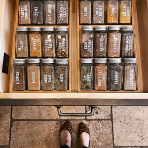 May include: A wooden drawer with a variety of spices in glass jars with white labels. The labels list the names of the spices, such as allspice, ancho, anise seed, basil, bay leaf, and more.