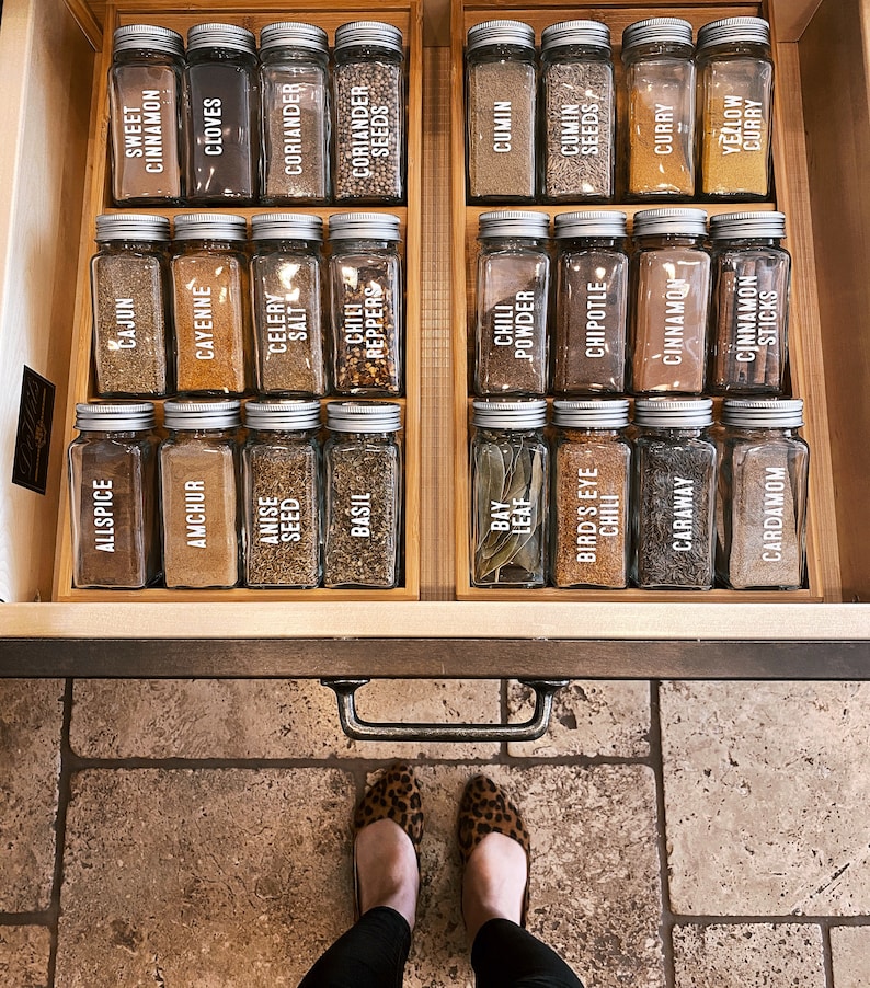 May include: A wooden drawer with a variety of spices in glass jars with white labels. The labels list the names of the spices, such as allspice, ancho, anise seed, basil, bay leaf, and more.