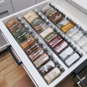 May include: A white drawer with clear glass jars filled with spices. The jars are labeled with the names of the spices, such as garlic, ginger, basil, and oregano. The drawer is open and the jars are visible.