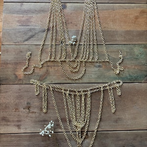 Op de afbeelding: Goudkleurige ketting body sieraden set, inclusief een top en een broekje. De kettingen zijn gerangschikt in een gedrapeerd ontwerp. De sieraden worden getoond op een houten oppervlak met een takje witte bloemen.