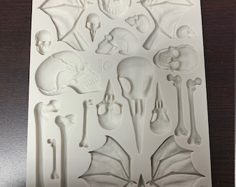 May include: White silicone mold with multiple cavities for making skull and bone shaped decorations. The mold features various skull designs, including a bird skull, a human skull, and a bat skull. There are also several bone shapes, including long bones and rib bones.