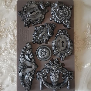 May include: A grey silicone mould featuring various ornate, antique-style decorative elements. Designs include keyholes, floral accents, and decorative hardware. The mould is designed for crafting and DIY projects.
