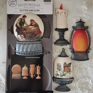 Könnte beinhalten: Eine Sammlung dekorativer Formen und fertiger Teile. Die Formen umfassen eine Krippenszene, eine Kerze, eine Laterne und eine Cloche mit Schmetterlingsdesign. Die Formverpackung ist 12,7 cm x 25,4 cm groß.