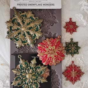 Könnte beinhalten: Eine Packung Dekorformen mit dem Text "FROSTED SNOWFLAKE WONDERS". Das Bild zeigt eine graue Form mit mehreren fertigen Schneeflocken-Ornamenten in Rot, Grün und Gold. Die Form misst 12.7cm x 25.4cm.