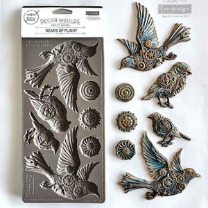 Könnte beinhalten: Eine graue Silikonform und mehrere dekorative Vogel- und Zahnradverzierungen. Die Form ist mit "DECOR MOULDS GEARS OF FLIGHT" beschriftet. Die Vögel und Zahnräder sind metallisch mit blauen und bronzefarbenen Akzenten.