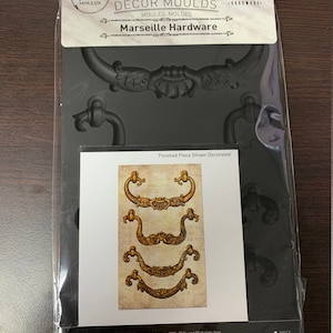 Puede incluir: Un molde de silicona negro para hacer herrajes decorativos. El molde mide 12,7 cm por 20,3 cm y presenta cuatro diseños diferentes de tiradores de puerta ornamentados. El molde es apto para alimentos y libre de BPA, PVC y ftalatos. El texto "Decor Moulds" y "Marseille Hardware" está impreso en el embalaje.