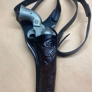 Può includere: Un revolver color argento in una fondina da spalla in pelle nera. La fondina presenta un motivo floreale marrone scuro. I cinturini in pelle nera sono regolabili. L'insieme è progettato per essere indossato sulla spalla.