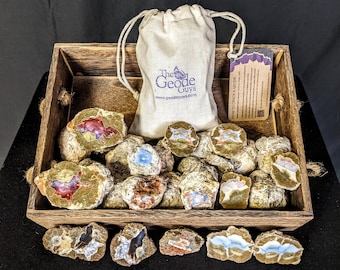 May include: A wooden crate filled with a variety of geode crystals. The geode crystals are various shades of brown, white, and blue. The crate is sitting on a black surface. The geode crystals are in a variety of shapes and sizes. The geode crystals are surrounded by a white drawstring bag with the text "The Geode Guys" printed on it. The bag is sitting on top of the geode crystals. A small tag is attached to the bag with the text "How to Crack a Geode" printed on it.