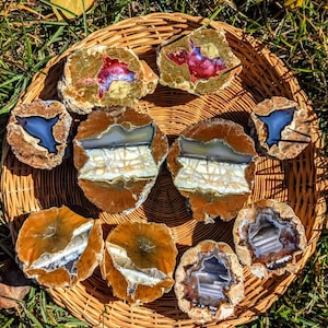 May include: A collection of polished geodes and stone slices arranged on a woven basket. The geodes display a variety of colors, including blue, red, and white, with a brown and tan exterior. The arrangement is set on a bed of green grass.