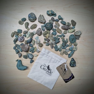 May include: A collection of small, green, blue, and brown rocks in a white drawstring bag with the text "The Geode Guys" printed on it. A brown tag with the same text is also included.