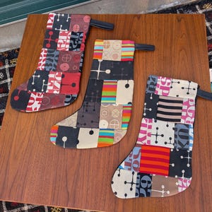 以下が含まれることがあります： 黒いハンドルが付いた3つのクリスマスストッキング。それぞれパッチワークのデザインが施されています。ストッキングは赤、黒、白、茶色など様々な色があります。ストッキングは茶色の木製の表面にあります。