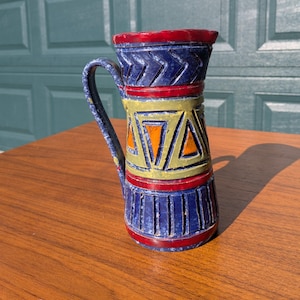May include: A decorative ceramic pitcher with a handle. The pitcher features a blue, red, yellow, and orange geometric design. The top and bottom rims are red, with a blue body and a yellow and orange center section.