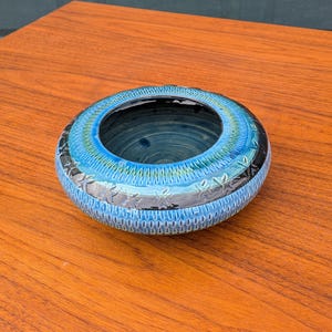Puede incluir: Un cuenco redondo de cerámica con una abertura central. El cuenco presenta un exterior azul turquesa texturizado, una banda negra y un interior azul oscuro. Se encuentra sobre una superficie de madera.