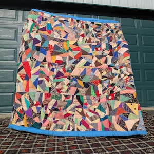 May include: A large rectangular quilt featuring a chaotic patchwork of various fabric scraps in diverse colors, patterns, and textures. The quilt is bordered by a solid bright blue fabric binding along the top and bottom edges.