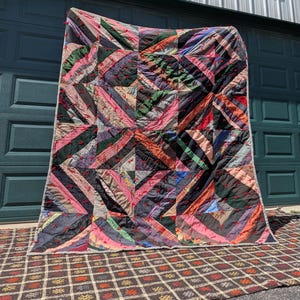 May include: A large, colorful quilt with a geometric pattern. The quilt features a patchwork design with various fabrics in shades of pink, black, green, and orange. Red stitching accents the diagonal lines, creating a striking visual effect.