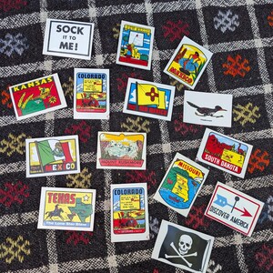 May include: A collection of vintage-style stickers featuring state names and imagery, including Kansas, Colorado, Texas, and New Mexico. One sticker reads "SOCK IT TO ME!" and another says "DISCOVER AMERICA". The stickers are arranged on a patterned fabric.