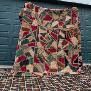 May include: A large quilt featuring a chaotic patchwork of irregular fabric shapes in shades of deep red, forest green, tan, and black. Each fabric piece is joined by thick, visible white zigzag stitching that creates a textured grid pattern.