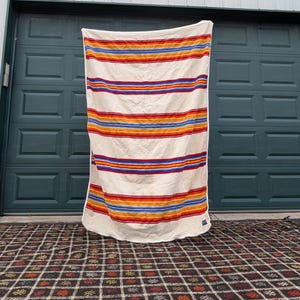May include: A cream-colored blanket with horizontal stripes in red, orange, yellow, and blue. The blanket is hanging in front of a dark green garage door. The blanket is approximately 70 inches long and 50 inches wide.
