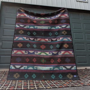 May include: A dark brown blanket with a Southwestern geometric pattern. The blanket features horizontal stripes in various colors, including teal, red, purple, and white. The design includes diamond and step-like shapes.