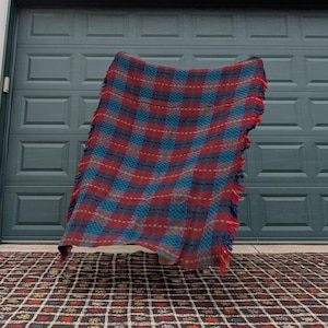 Puede incluir: Una manta vintage a cuadros en tonos rojos, azules y verdes con bordes con flecos. La manta está drapeada, mostrando su patrón tejido. El fondo presenta una puerta de garaje verde y una alfombra estampada.