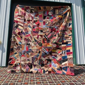 Puede incluir: Una colcha rectangular grande de estilo crazy quilt, construida con una multitud de retazos de tela irregulares en diversos colores, texturas y patrones. El textil presenta intrincadas costuras decorativas a lo largo de las uniones.