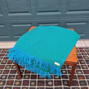 May include: A teal and green woven blanket with a geometric pattern and blue fringe. The blanket is draped over a small wooden table. The background includes a teal garage door and a patterned rug.