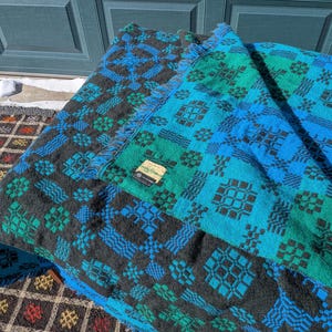 Op de afbeelding: Een opgevouwen deken met een geometrisch patroon in zwart, blauwgroen en blauw. De deken heeft een gefranjerde rand en een klein label. Het ontwerp heeft herhaalde vierkante en ruitvormige vormen.