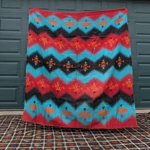May include: A vibrant blanket with a Southwestern design. The blanket features a repeating pattern of zigzags and geometric shapes in red, turquoise, black, and orange. The blanket is displayed against a neutral background.