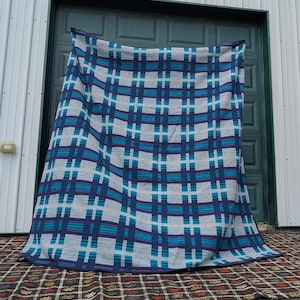 May include: A large rectangular blanket featuring a repeating plaid pattern in shades of light gray, dark navy blue, bright turquoise, and magenta. The textile displays a grid of intersecting horizontal and vertical stripes across the entire surface.