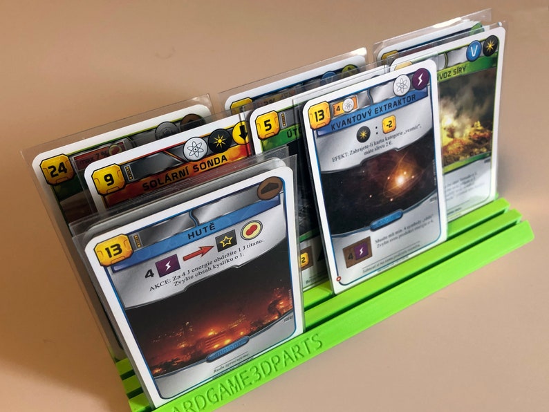 May include: A green card game organizer with six clear plastic card holders. The card holders are filled with cards featuring colorful illustrations and text in a foreign language. The text on the cards includes numbers, symbols, and the words "Solarni Sonda", "Hute", "Kvantovy Extraktor", and "Voz Siry".