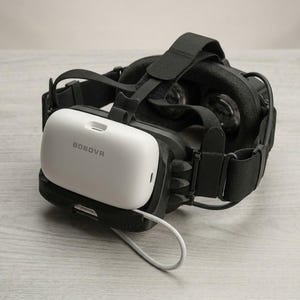 Pode incluir: Um headset de realidade virtual preto com uma bateria branca acoplada na parte traseira, com a marca BOBOVR impressa em cinza na superfície branca. O dispositivo inclui tiras de tecido preto ajustáveis e um cabo de carregamento branco.