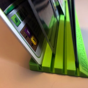 May include: A green plastic card holder with five slots for holding cards upright. The holder has the word "BOARDGAME" printed on the side.