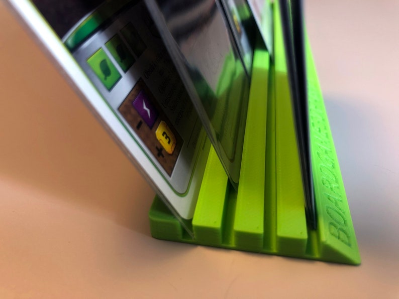May include: A green plastic card holder with five slots for holding cards upright. The holder has the word "BOARDGAME" printed on the side.