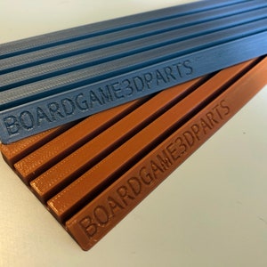 May include: Two 3D printed organizers for board games. The top organizer is blue and the bottom organizer is brown. Both organizers have the text "BOARDGAME3DPARTS" printed on them.