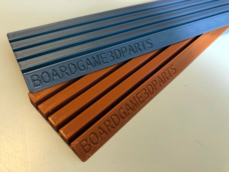 May include: Two 3D printed organizers for board games. The top organizer is blue and the bottom organizer is brown. Both organizers have the text "BOARDGAME3DPARTS" printed on them.