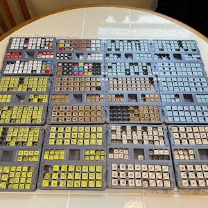 Peut inclure: Une collection de plateaux de rangement en plastique gris remplis de nombreux petits dés carrés de différentes couleurs, notamment jaune, bleu clair, beige et blanc. Chaque dé présente des symboles ou des chiffres imprimés, organisés dans les compartiments.