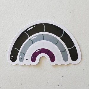 May include: A black, grey, and purple rainbow sticker with a white background. The rainbow is made up of three curved lines, each with a different colour. The top line is black, the middle line is grey, and the bottom line is purple.